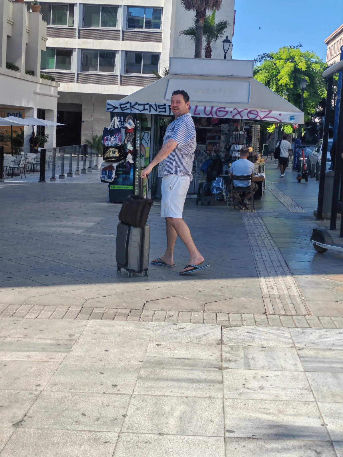 A traveller walking with a wheeled suitcase past a street kiosk in central Athens on a sunny morning