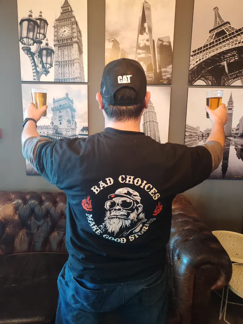 A traveller in a 'Bad Choices Make Good Stories' shirt raises two beers toward framed photos of world cities