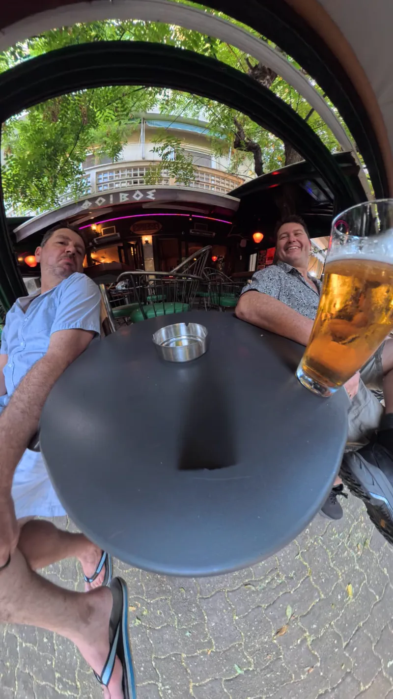 Two friends laughing over a cold beer at Foivos bar in a leafy Athens neighbourhood square
