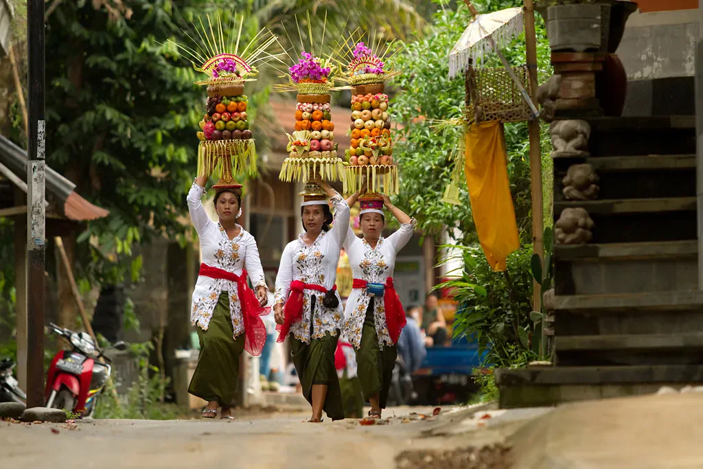 Balinese women in temple dress carrying tall fruit offerings on their heads along a village street