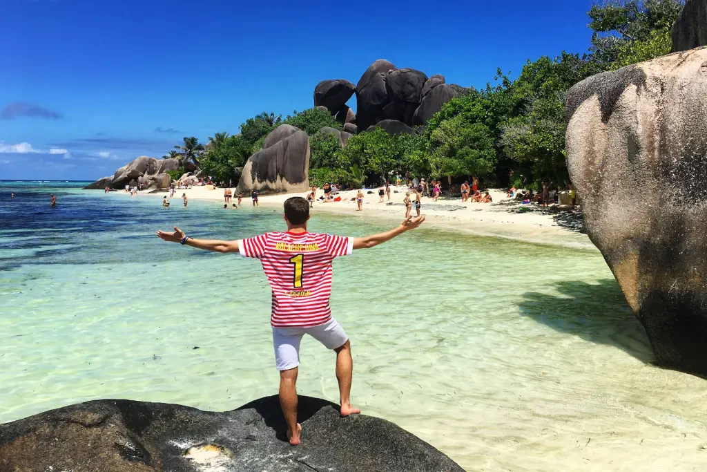 Traveller with arms outstretched on a granite rock at Anse Source d'Argent, La Digue, with giant boulders and shallow turquoise water