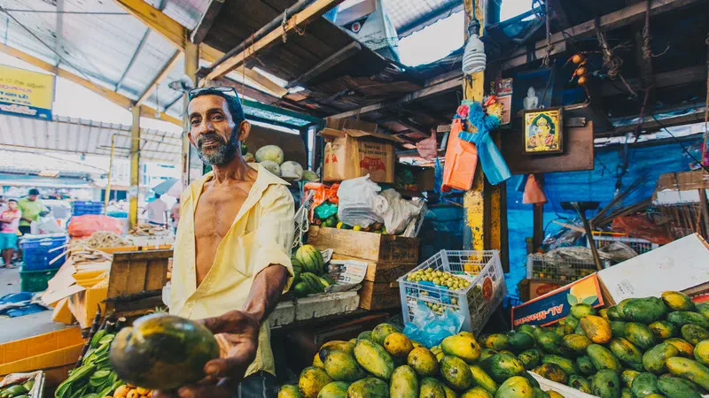 Smiling fruit vendor offering a papaya at a colourful market stall in Sri Lanka