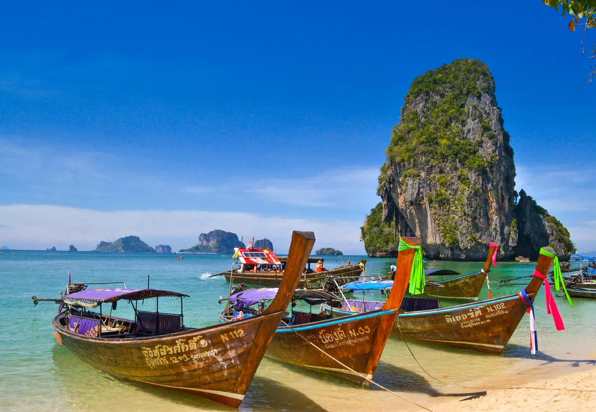 Colourful longtail boats on the beach at Railay, Krabi, with a limestone karst rising from turquoise water in Thailand